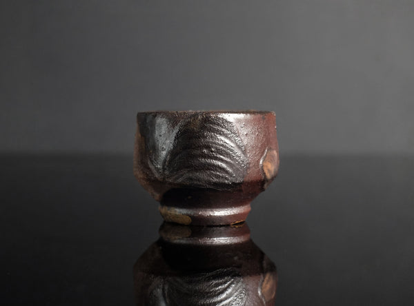 A small handmade ceramic cup with a textured, rustic finish in dark brown and black tones, set against a dark gray background with a soft reflection below.