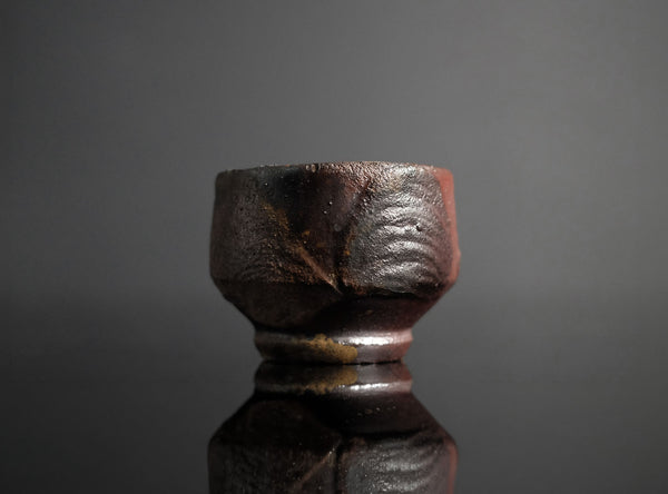 A small handmade ceramic cup with a textured, rustic finish in dark brown and black tones, set against a dark gray background with a soft reflection below.