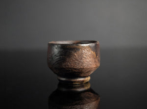 A small handmade ceramic cup with a textured, rustic finish in dark brown and black tones, set against a dark gray background with a soft reflection below.