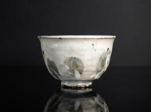 A vintage ceramic bowl with a white glaze, featuring irregular gray and brown patches and speckles, set against a dark reflective background.