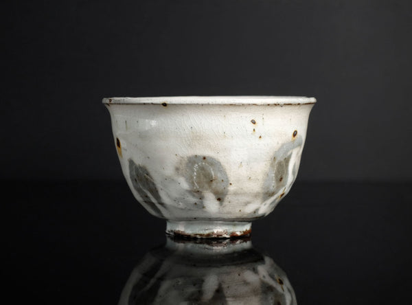 A vintage ceramic bowl with a white glaze, featuring irregular gray and brown patches and speckles, set against a dark reflective background.
