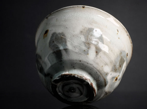 A vintage ceramic bowl with a white glaze, featuring irregular gray and brown patches and speckles, set against a dark reflective background.