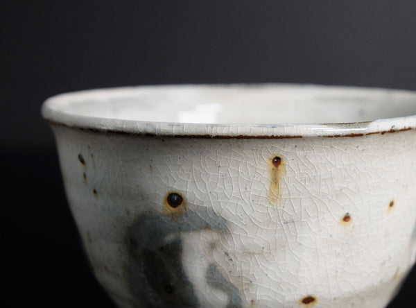 A close-up of a ceramic bowl with a textured off-white glaze, featuring a crackled finish, small brown specks, and a dark gray painted area on the front, set against a dark background.