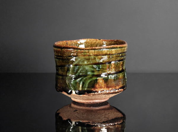 A handmade ceramic tea bowl with a glossy, textured glaze in earthy brown and green tones, displayed on a reflective black surface against a dark gray background.