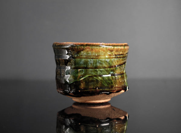 A handmade ceramic tea bowl with a glossy, textured glaze in earthy brown and green tones, displayed on a reflective black surface against a dark gray background.