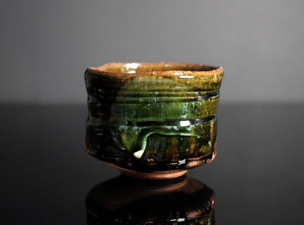 A handmade ceramic tea bowl with a glossy, textured glaze in earthy brown and green tones, displayed on a reflective black surface against a dark gray background.