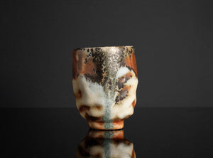 A handmade ceramic cup with a textured, organic glaze pattern in brown, cream, and black, standing on a reflective black surface against a dark gray background.