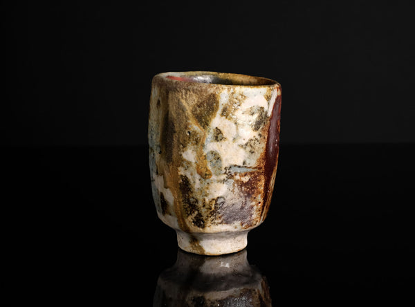 A handmade ceramic cup with a textured, mottled glaze in earthy brown, beige, and white tones, standing on a reflective black surface against a solid black background.