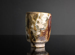 A handmade ceramic cup with a textured, mottled glaze in earthy tones of brown, beige, and white, standing on a reflective black surface against a dark gray background.