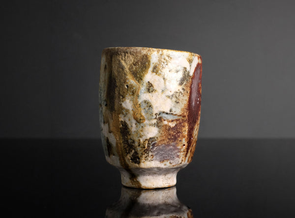 A handmade ceramic cup with a textured, mottled glaze in earthy tones of brown, beige, and white, standing on a reflective black surface against a dark gray background.