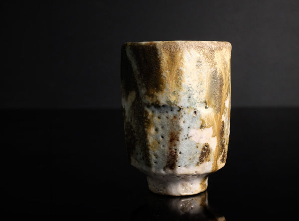 A handmade ceramic cup with a textured, rustic finish featuring a mix of brown, beige, and white glazes, set against a dark black background.