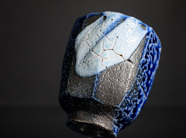 A handmade ceramic cup with a textured, faceted design, featuring a glossy blue and matte black glaze with a light blue and white cracked tile motif on the front, set against a dark background.