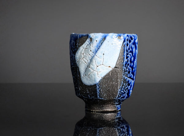 A handmade ceramic cup with a textured, faceted design, featuring a glossy blue and matte black glaze with a light blue and white cracked tile motif on the front, set against a dark background