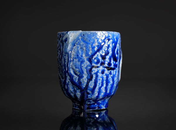 A handmade ceramic cup with a textured, faceted design, featuring a glossy blue and matte black glaze with a light blue and white cracked tile motif on the front, set against a dark background
