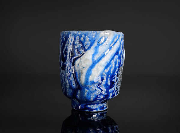 A small ceramic cup with a blue and white marbled, tree-like glaze pattern, shown on a dark reflective surface against a black background.