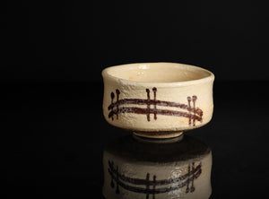 A handmade ceramic matcha bowl with a textured cream glaze and dark brown brushstroke patterns, displayed on a reflective black surface against a solid black background.