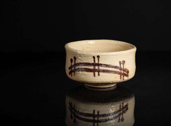 A handmade ceramic matcha bowl with a textured cream glaze and dark brown brushstroke patterns, displayed on a reflective black surface against a solid black background.