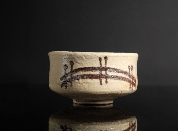 A handmade ceramic tea bowl with a textured off-white glaze and dark brown painted brushstrokes, set against a dark black background with a soft reflection below.