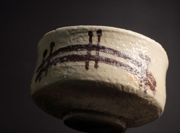 A close-up of a handmade ceramic bowl with a textured off-white glaze and dark brown painted brushstrokes, set against a dark background.