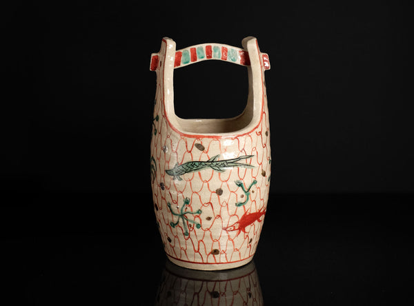 A ceramic pitcher with a loop handle, decorated with a red and green fish pattern on a white background, set against a solid black backdrop.