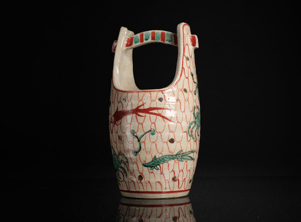 A ceramic pitcher with a loop handle, decorated with a red and green fish pattern on a white background, set against a solid black backdrop.
