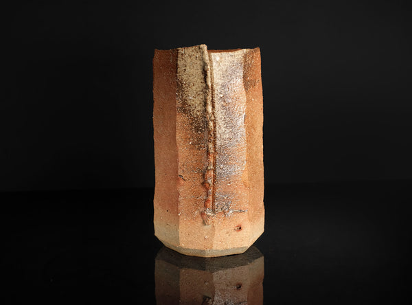 A handmade ceramic vase with a textured, geometric, faceted design in earthy terracotta and beige tones, standing on a reflective black surface against a solid black background.