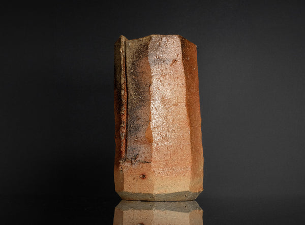 A handmade ceramic hexagonal vase with a rustic, textured surface in earthy terracotta and brown tones, standing on a reflective black surface against a dark background.