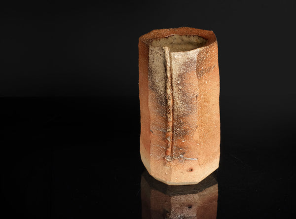 A handmade ceramic vase with a textured, rustic finish in earthy terracotta and brown tones, standing on a reflective black surface against a black background.