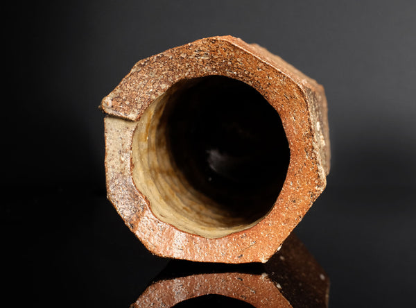 A close-up of a hexagonal ceramic tile or tile fragment with a rustic, textured brown and orange-brown glaze, set against a dark reflective background.