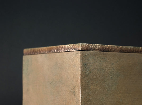 Close-up of a square metal block corner showing a textured, hammered metallic surface in a warm bronze or copper tone against a dark background.