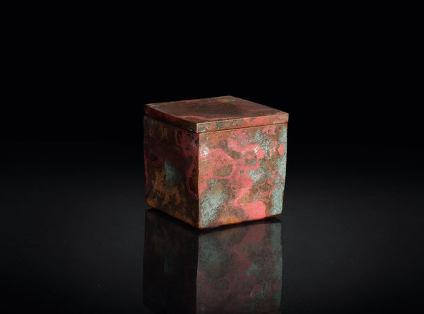 A small square decorative box with a lid, featuring a textured metallic surface in coppery red and green patina tones, set against a solid black background with a soft reflection below.