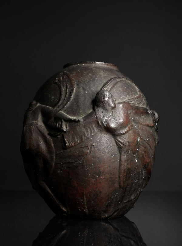 A dark metallic sculpture or decorative vase featuring a stylized human figure in relief, photographed against a dark studio background with a reflective surface below.