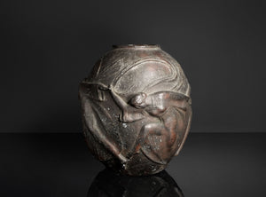 A dark metallic sculpture or decorative vase featuring a stylized human figure in relief, photographed against a dark studio background with a reflective surface below.