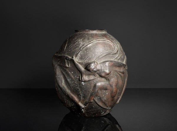 A dark metallic sculpture or decorative vase featuring a stylized human figure in relief, photographed against a dark studio background with a reflective surface below.