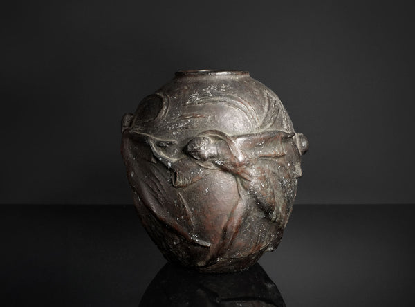 A dark metallic sculpture or decorative vase featuring a stylized human figure in relief, photographed against a dark studio background with a reflective surface below.