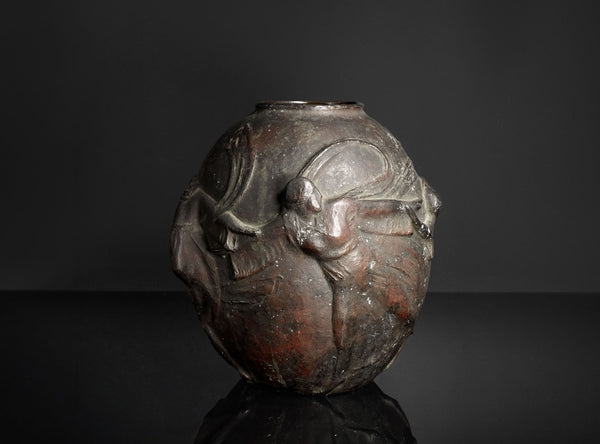 A dark metallic sculpture or decorative vase featuring a stylized human figure in relief, photographed against a dark studio background with a reflective surface below.