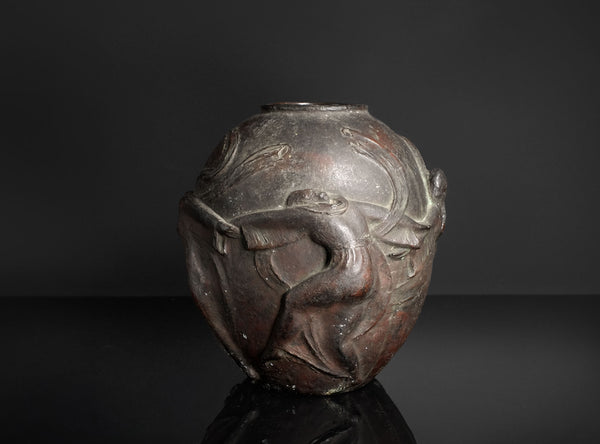 A dark metallic sculpture or decorative vase featuring a stylized human figure in relief, photographed against a dark studio background with a reflective surface below.