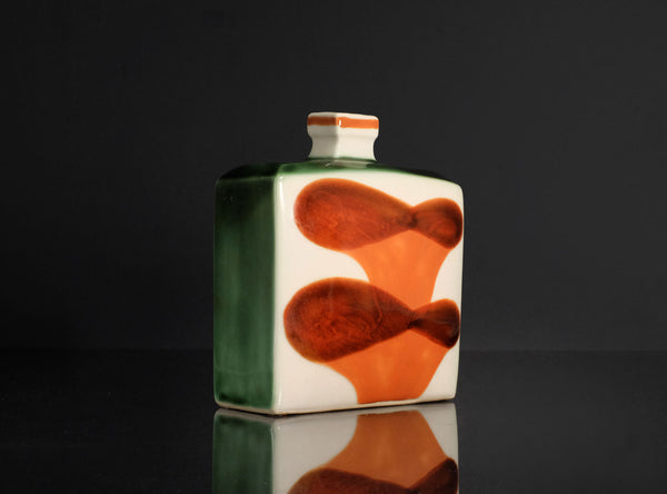 A square ceramic bottle with a small neck and a stopper, featuring a white body with two large orange abstract shapes and a dark green side panel, set against a dark reflective background.