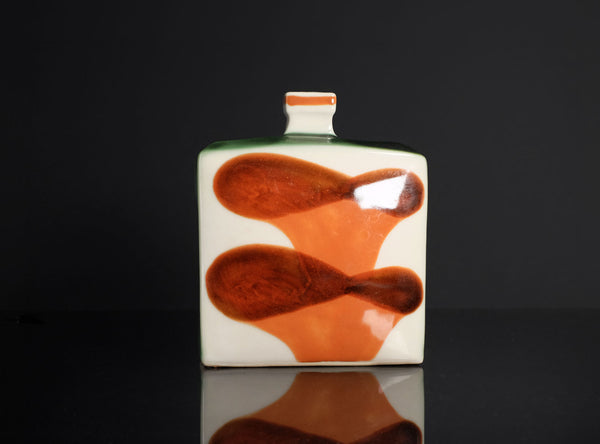A square white ceramic vase with a glossy finish, featuring two abstract organic shapes in orange-brown and a small green band near the top, set against a dark reflective background.