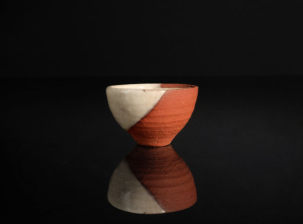 A handmade ceramic cup with a diagonal split design of terracotta orange and cream, set against a solid black background with a clear reflection below.