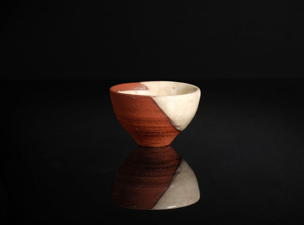 A handmade ceramic cup with a diagonal split design, featuring a reddish-brown textured clay exterior and a smooth off-white glazed interior, set against a solid black background with a clear reflection below.
