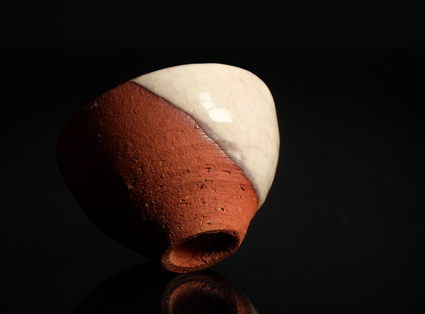 A handmade ceramic cup with a dual-tone finish, featuring a textured terracotta red lower half and a glossy off-white glazed upper half, set against a solid black background.