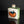 Load image into Gallery viewer, A small square ceramic bottle or tumbler with a narrow neck, featuring a hand-painted design of an orange with a blue leaf on a white body and green glazed sides, set against a dark reflective background.
