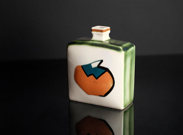 A small square ceramic bottle or tumbler with a narrow neck, featuring a hand-painted design of an orange with a blue leaf on a white body and green glazed sides, set against a dark reflective background.