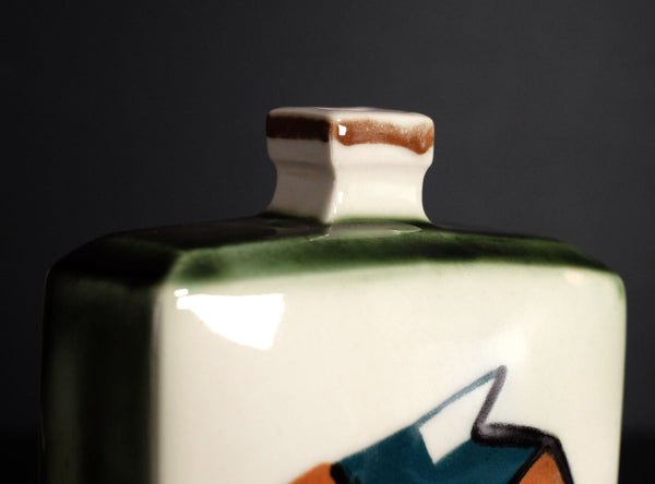 Close-up of a glossy ceramic bottle or vase with a white body, dark green band, and abstract blue, orange, and black painted details near the bottom, set against a dark gray background.