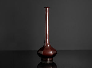 A glossy dark reddish-brown ceramic vase with a long, slender neck and a rounded base, displayed on a reflective black surface against a dark gray background.