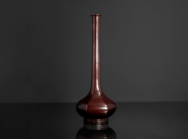 A glossy dark reddish-brown ceramic vase with a long, slender neck and a rounded base, displayed on a reflective black surface against a dark gray background.