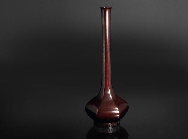 A glossy dark reddish-brown ceramic vase with a long, slender neck and a rounded base, displayed on a reflective black surface against a dark gray background.