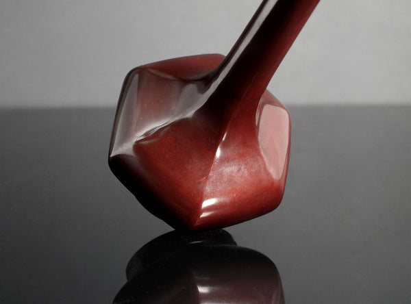 A close-up studio shot of a glossy, faceted sculptural object in a deep reddish-brown color, resting on a reflective dark surface against a neutral gray background.