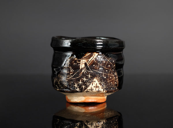 A small handmade ceramic bowl with a glossy black glaze, featuring hand-painted gold and white starry patterns on the front, resting on a reflective black surface against a dark gray background.